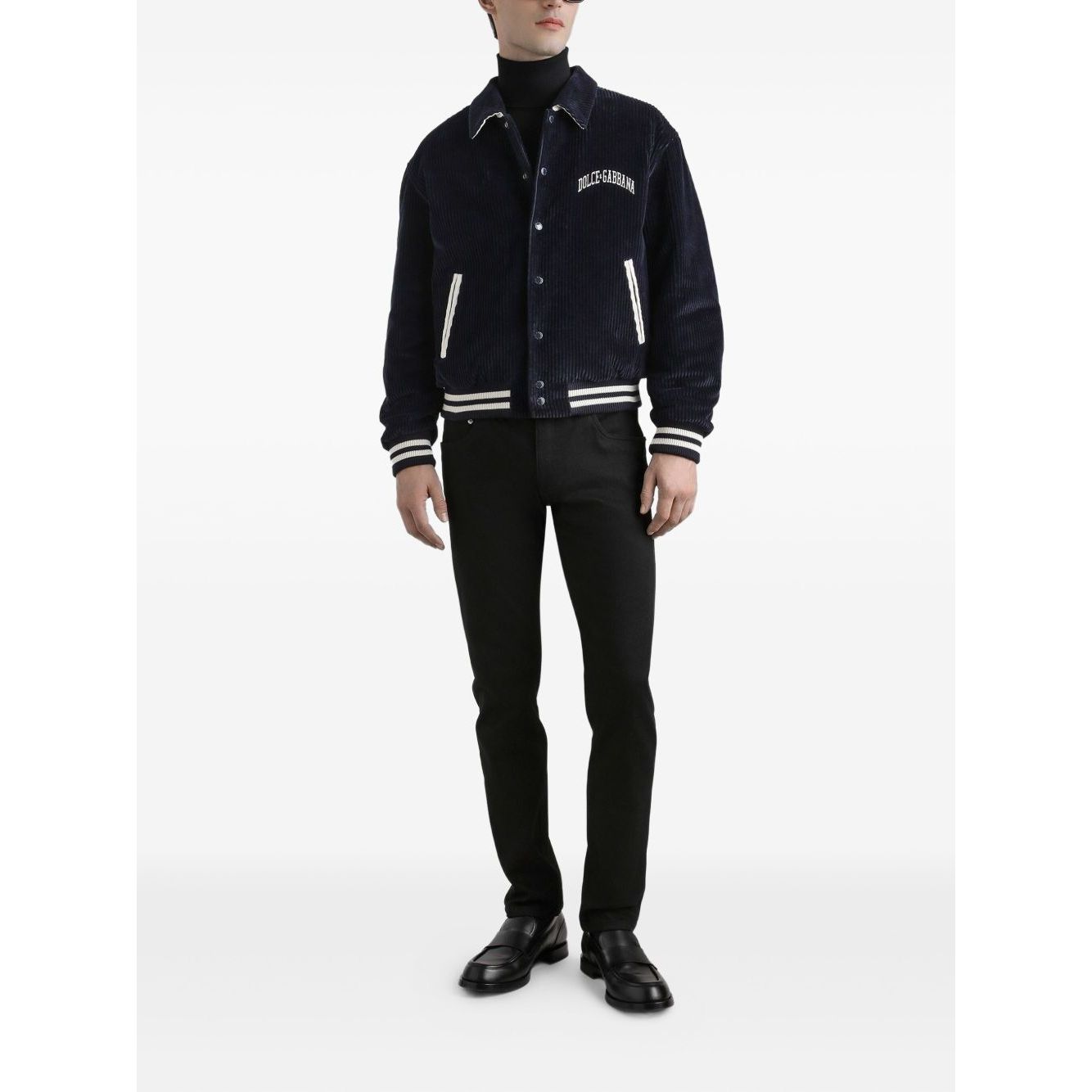 Man wearing Dolce & Gabbana five pockets logo plaque Denim cotton jeans and navy jacket with white stripes, standing against white background.