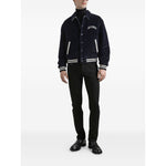 Man wearing Dolce & Gabbana five pockets logo plaque Denim cotton jeans and navy jacket with white stripes, standing against white background.